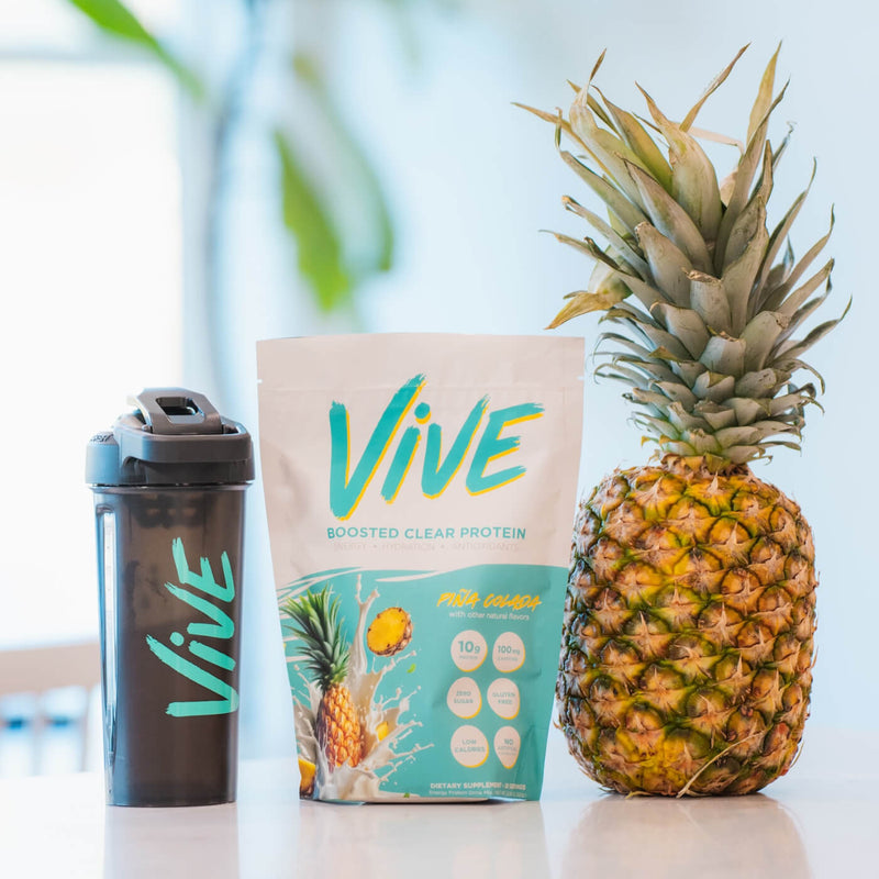 Vive protein package, shaker, and pineapple on a light surface with blurred greenery in the background